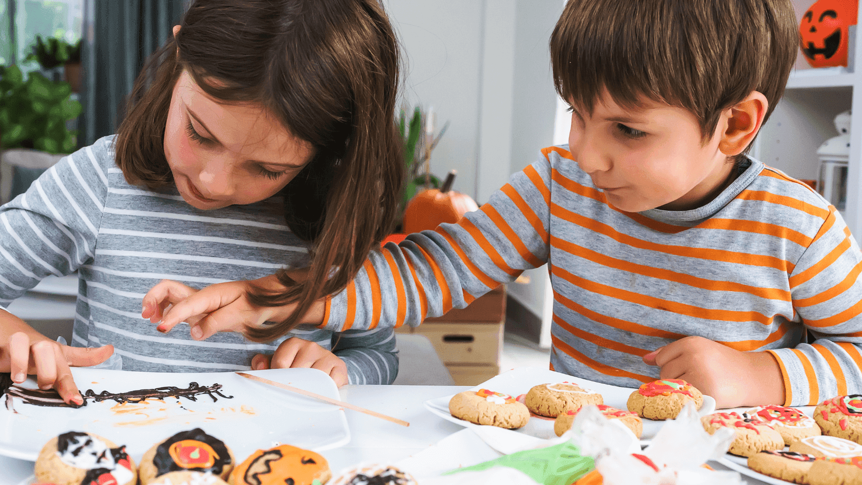 Two children decorate Halloween cookies with frosting and chocolate at a dining table.