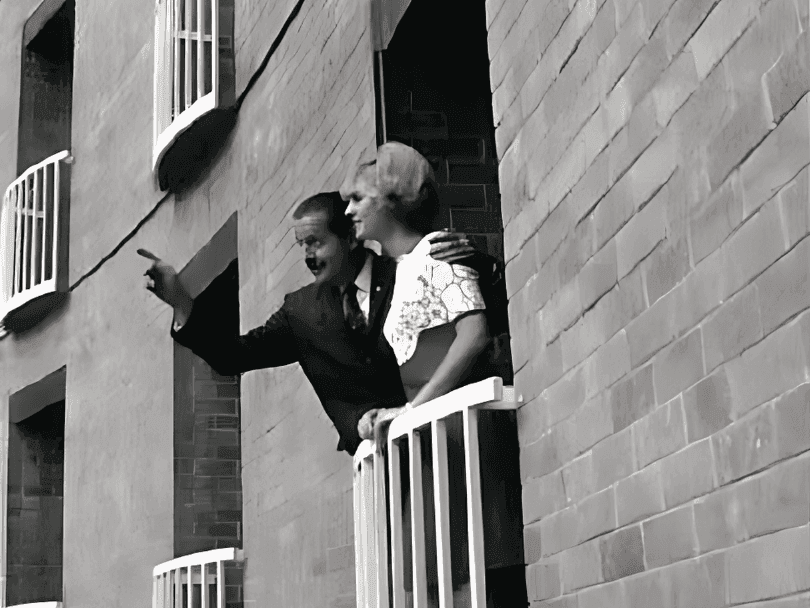 Black and white photograph of couple embracing on building balcony with geometric brick architecture