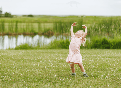 Young girl in a pink dress dancing with raised arms on a green lawn by a tranquil pond outdoor area at Rodd Hotels & Resorts