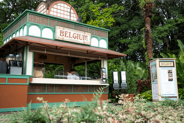 A green and burnt sienna colored food pavilion with a sign that reads BELGIUM, surrounded by greenery.