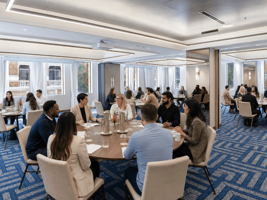 Professionals engaged in meetings around tables in a bright conference room with blue patterned carpet and large windows.