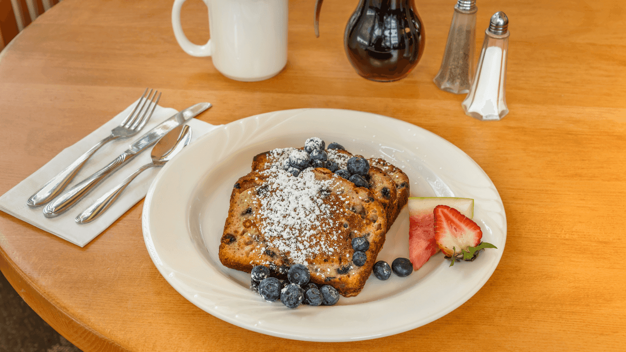 A plate of French toast with blueberries, strawberries, and powdered sugar on a wooden table with silverware and a mug.