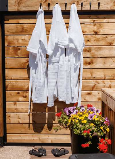 White waffle robes hanging from hooks on a wood fence above flip-flops by a colorful floral planter at Rodd Moncton
