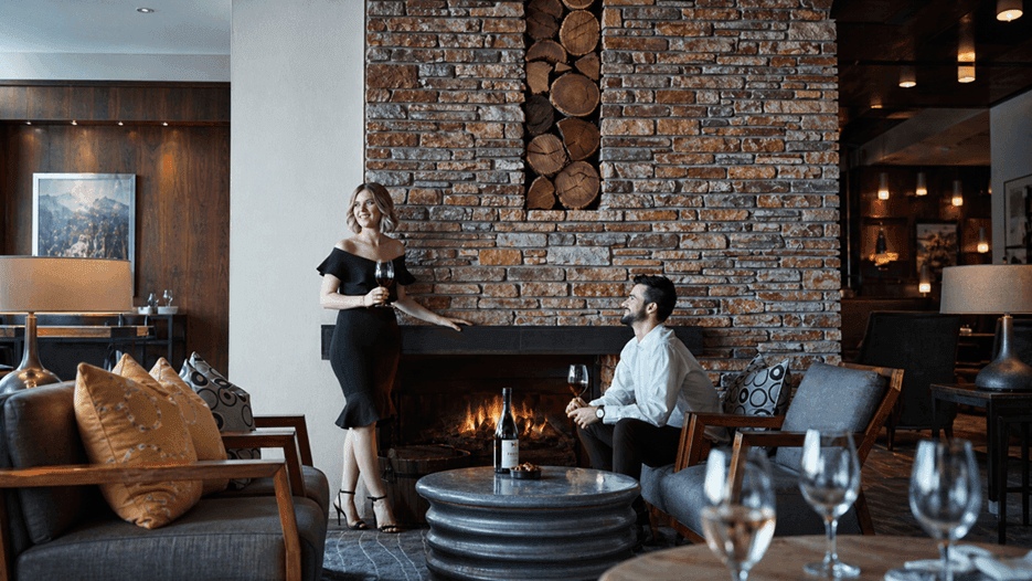 Couple enjoying wine in a cozy lounge with fireplace under offer Make Yourself at Home with Extended Winter Stays.