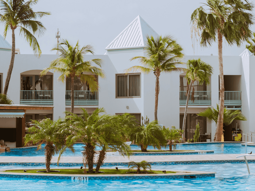 Palm trees by a blue swimming pool surrounding white buildings under a sunny sky at The Mill Resort & Suites Aruba