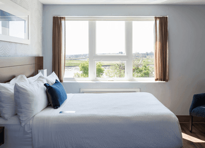 Comfortable bed with blue accent pillow near a large bright window overlooking lush green nature at Rodd Moncton