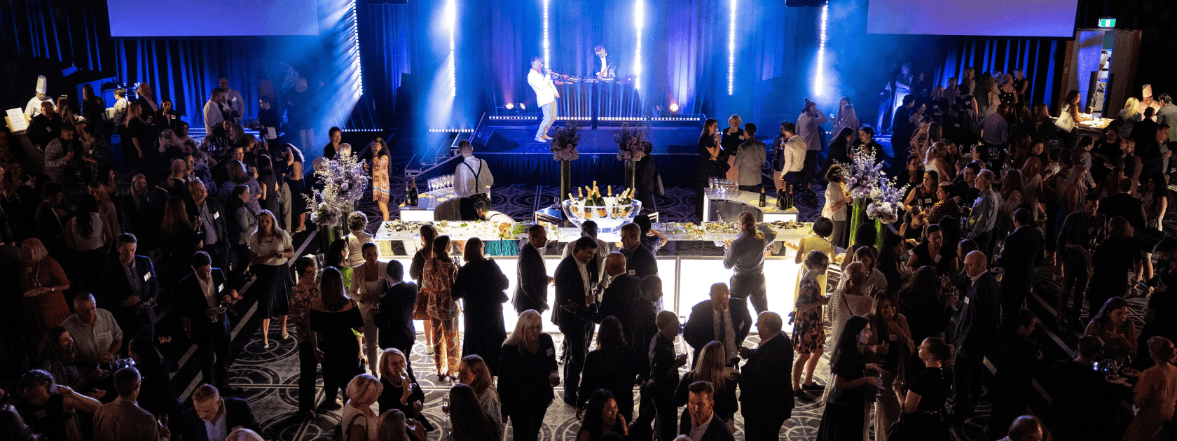 Large group of people gathered at a formal event with performers on stage and illuminated by blue lights.