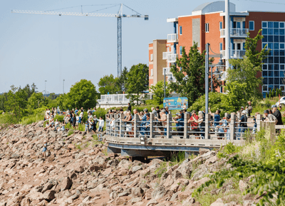 Crowds gathered on a scenic riverside trail platform on a sunny day beside the waterfront trail near Rodd Moncton