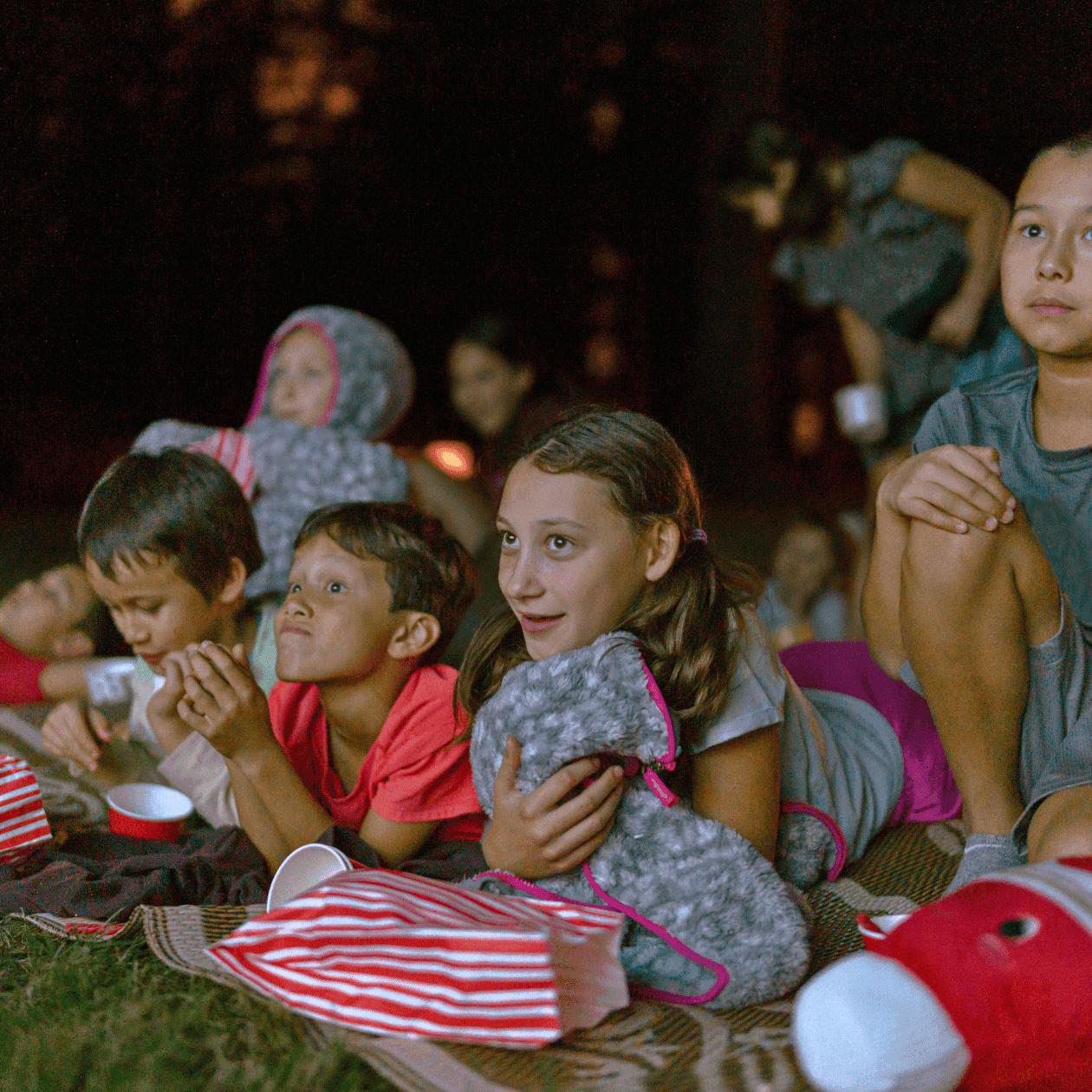 Children sitting on blankets in the dark, with snacks and stuffed animals, watching a movie outdoors.