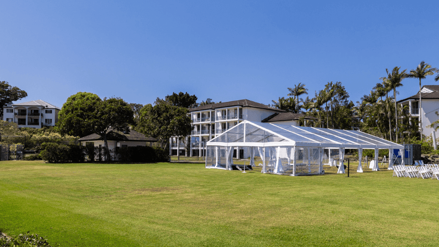 Large tent and chairs on a green lawn in front of buildings and palm trees under a clear blue sky.