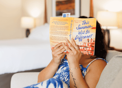Woman lounging on a sofa and reading a novel while relaxing in a cozy, bright guest room setting at Rodd Hotels & Resorts