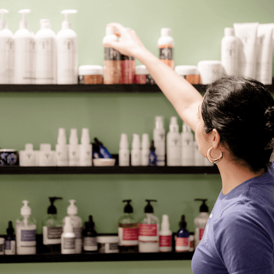 Staff member reaching for skincare products lined across dark wood shelves on a green wall at La Tourelle Inn & Spa