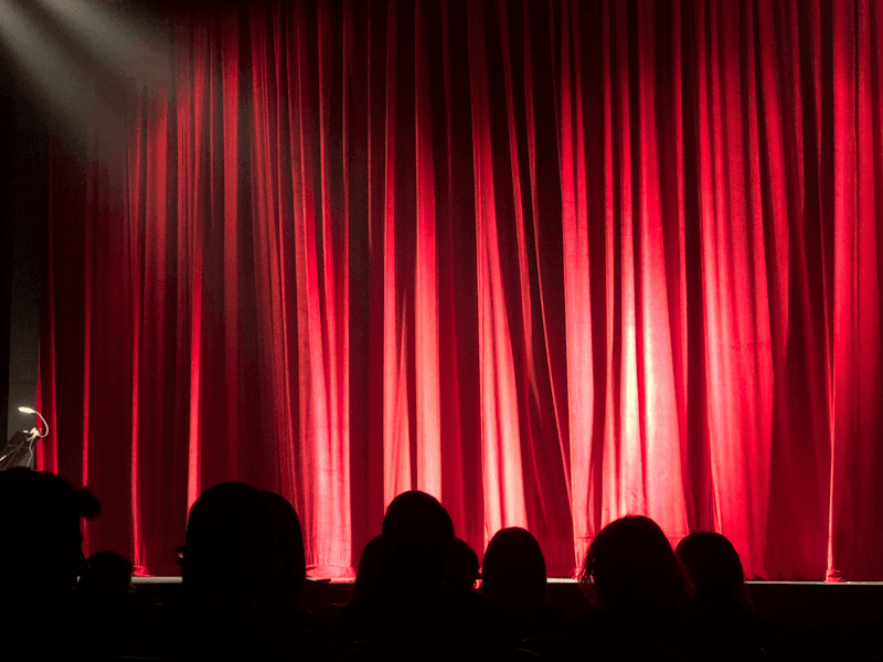 Red theatre curtains illuminated by stage lights with audience silhouettes waiting for a performance.