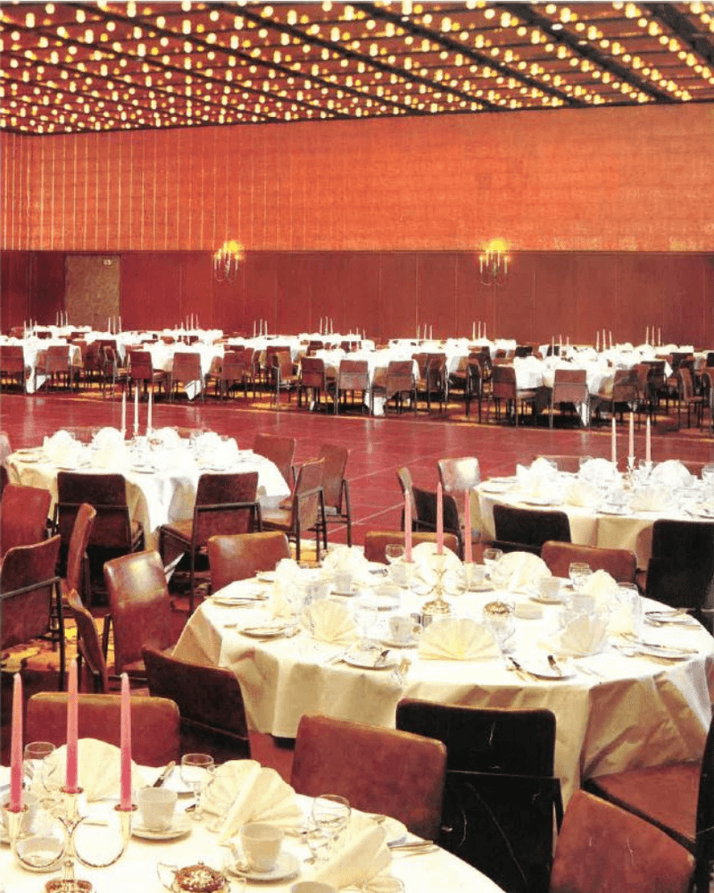 Banquet hall with numerous tables set with white tablecloths, chairs, and decorative lighting fixtures.