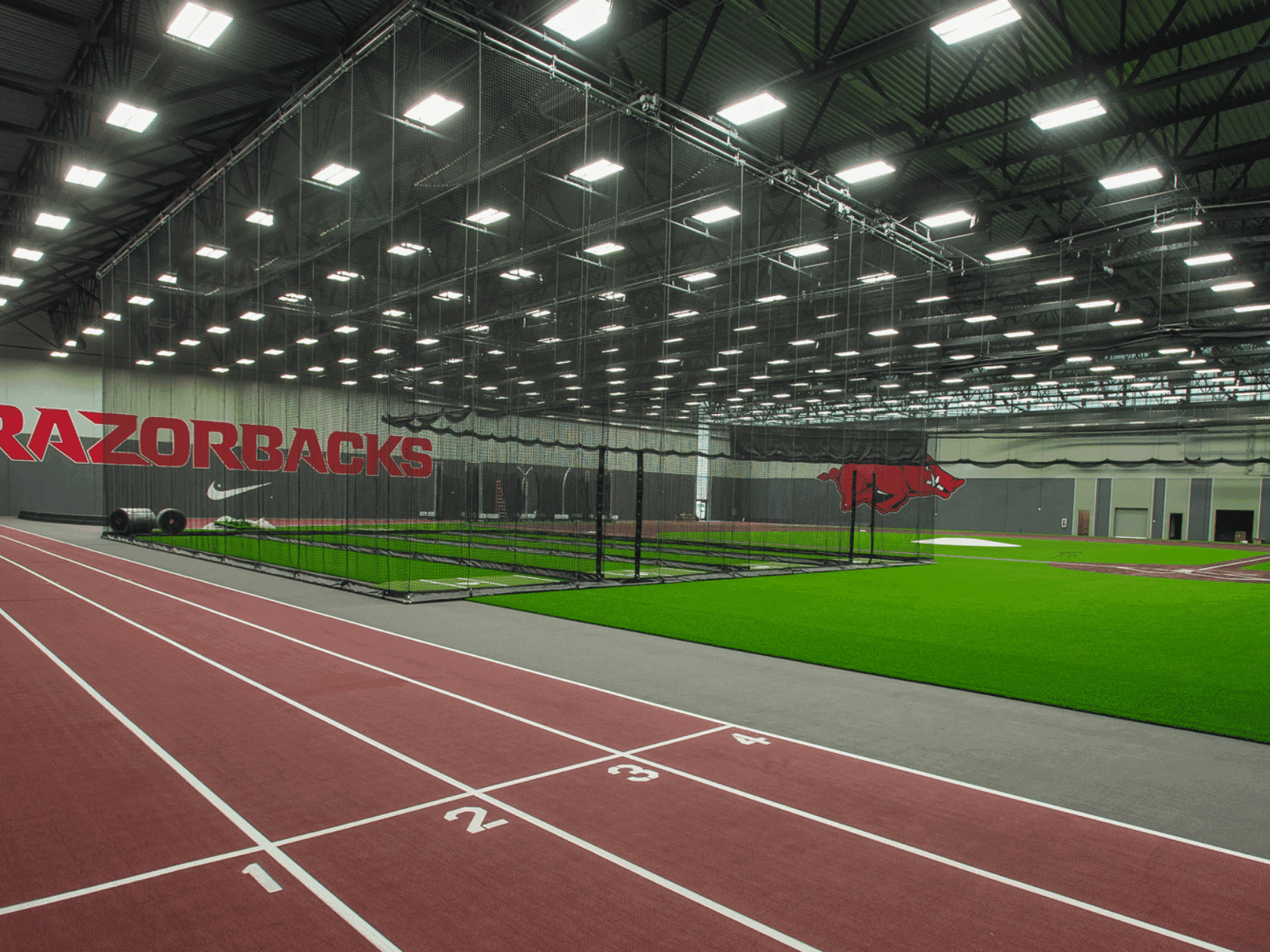 Indoor sports facility with red track, numbered lanes, and a green field, under a metal-framed ceiling.