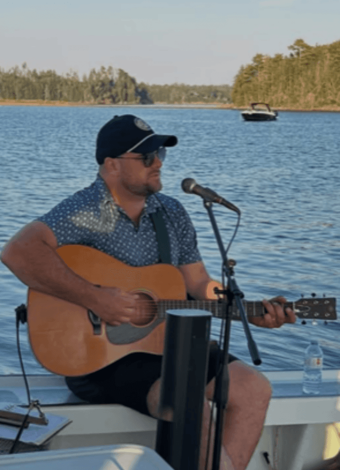 Man wearing cap and sunglasses playing guitar on a boat in front of a scenic river at Rodd Brudenell River Resort