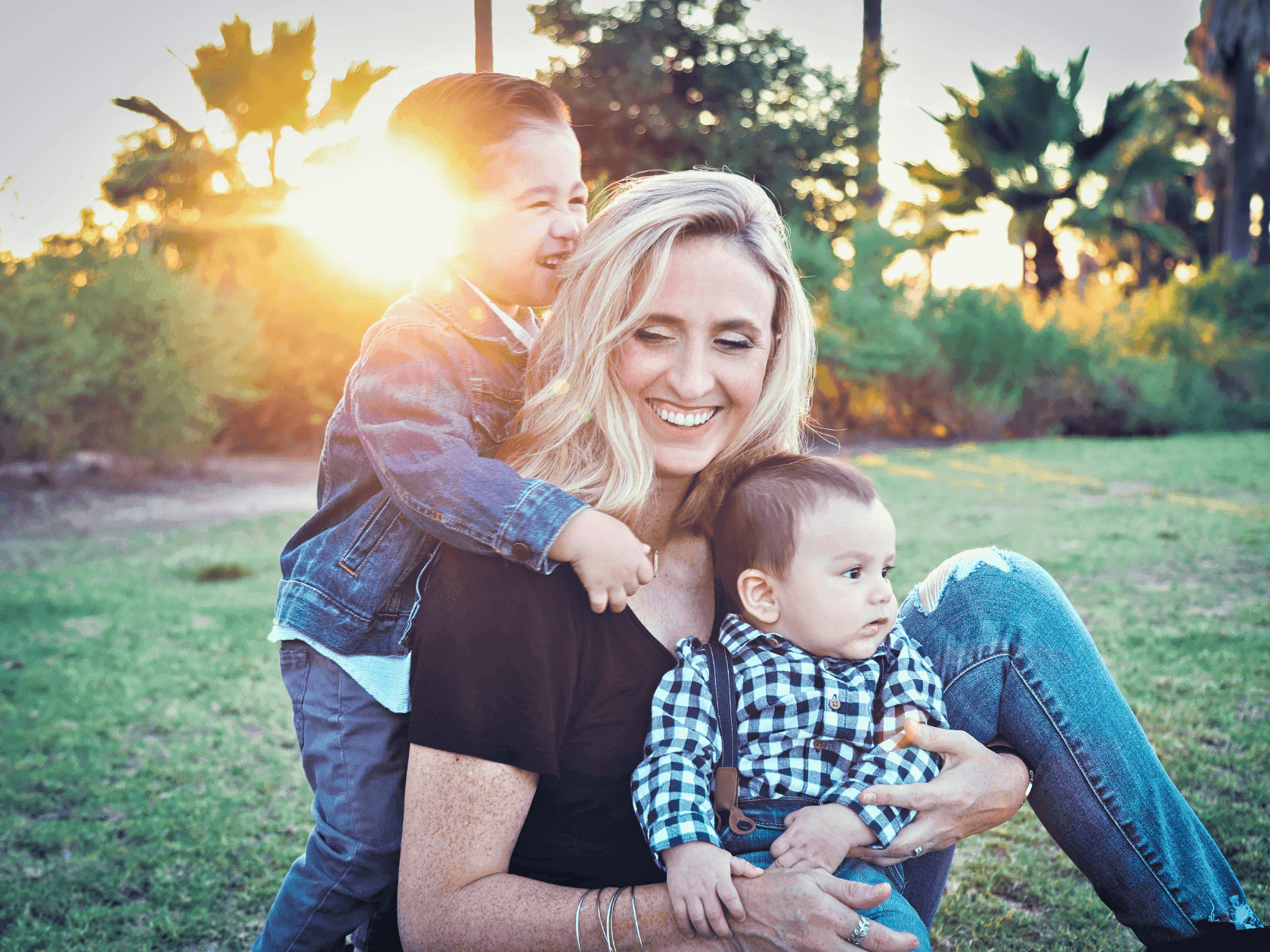 Mother with blonde hair holding two children in park with palm trees and sunset in background.