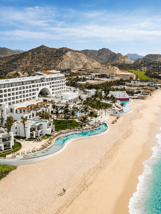 Aerial view of a beachfront resort with pools, palm trees, and turquoise ocean waters along a sandy shore.