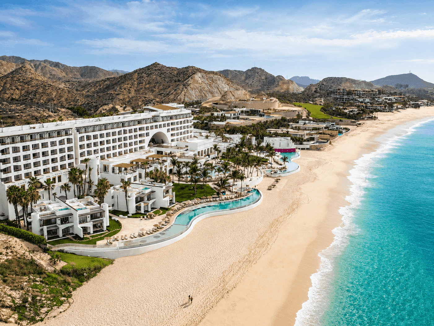 Vista aérea de resort costero con piscinas serpenteantes, playa de arena dorada y montañas al fondo bajo cielo azul