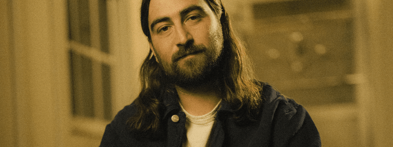Man with long hair and beard standing indoors for Noah Kahan's The Great Divide Tour 2026.