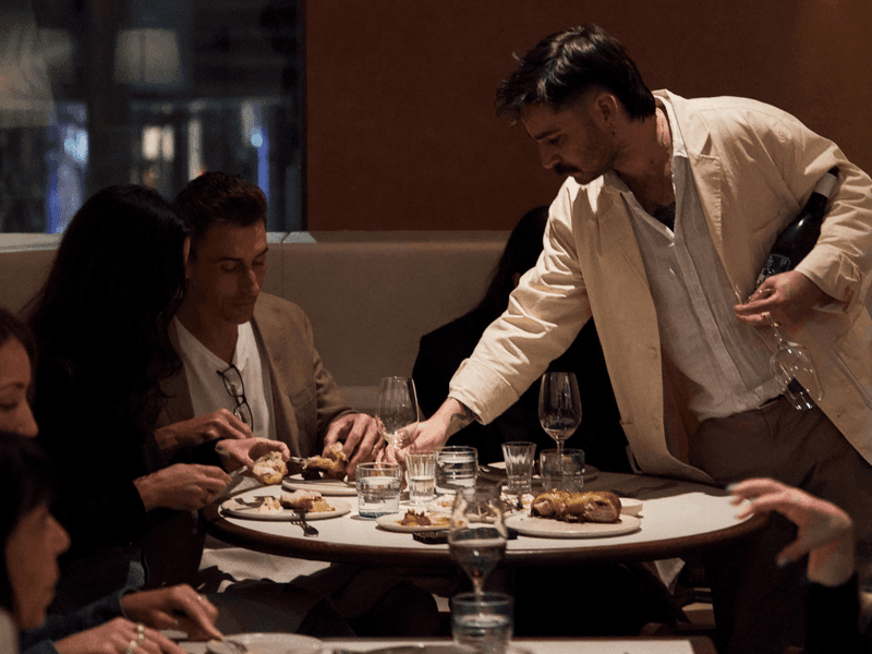 A waiter holds a wine bottle over a table of diners in a dimly lit restaurant.
