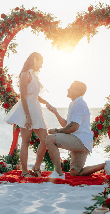 A man is kneeling and proposing to a woman under a heart-shaped floral arch on the beach.