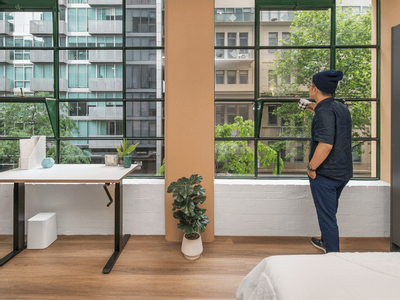 Man standing by windows with city view, holding a cup, near a desk and a bed.
