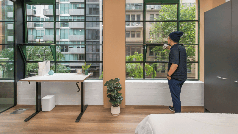 Man standing by windows with city view, holding a cup, near a desk and a bed.