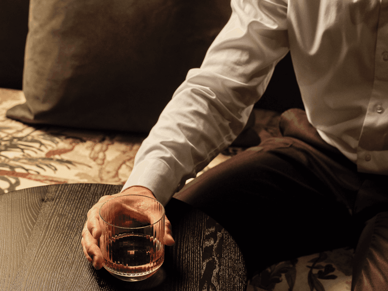 Cozy lounge scene with a person holding a glass of whiskey on a dark table, surrounded by plush seating.
