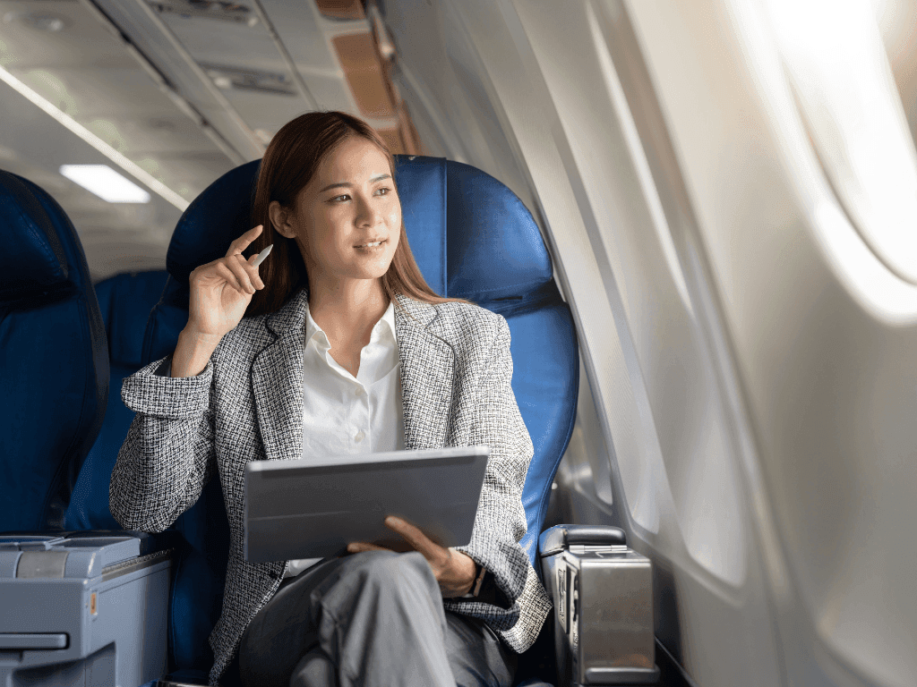 Woman in business attire sitting on an airplane, holding a tablet, and talking on the phone.