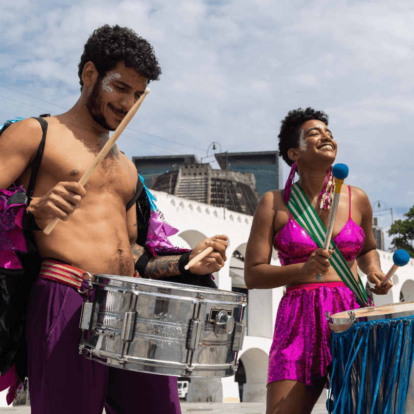 A male and female performer in vibrant, festive costumes energetically playing drums during a lively celebration.