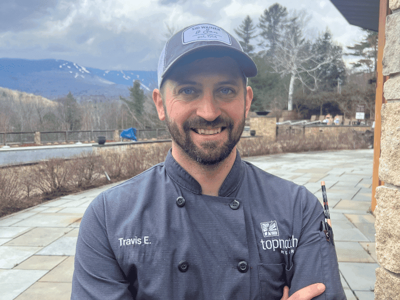Chef Travis smiling with arms crossed on a stone patio with scenic view.
