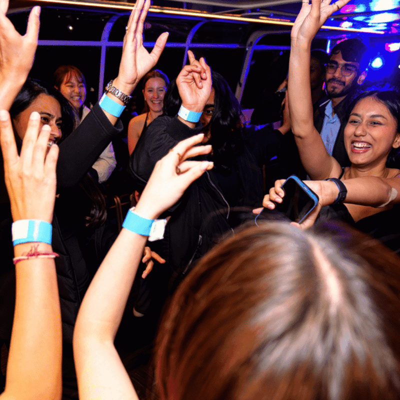 Group of young people dancing with raised arms and blue wristbands at a vibrant nighttime event with colorful lighting.