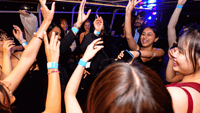 Group of young people dancing with raised arms and blue wristbands at a vibrant nighttime event with colorful lighting.