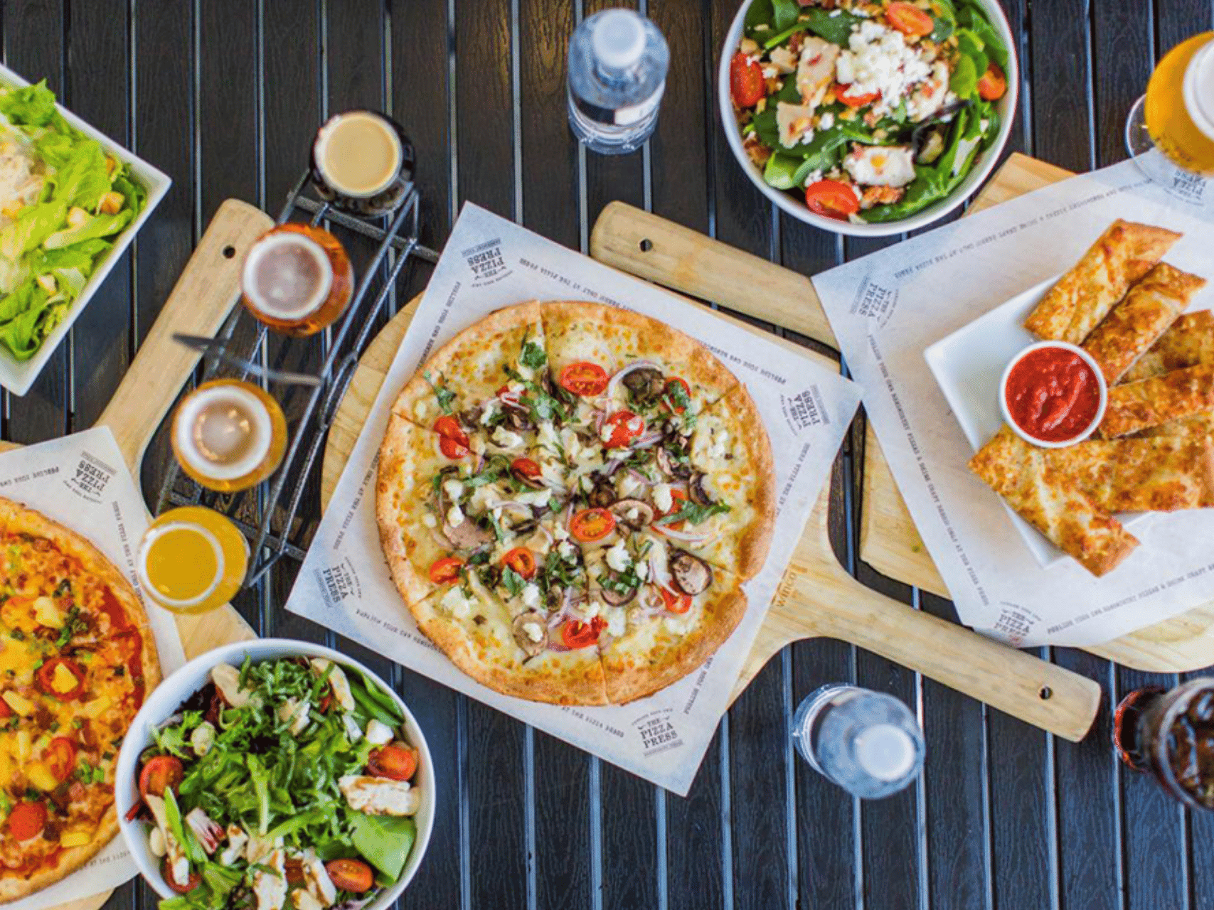 Overhead view of pizzas, salads, and beverages on a wooden table at The Pizza Press at The Anaheim Hotel