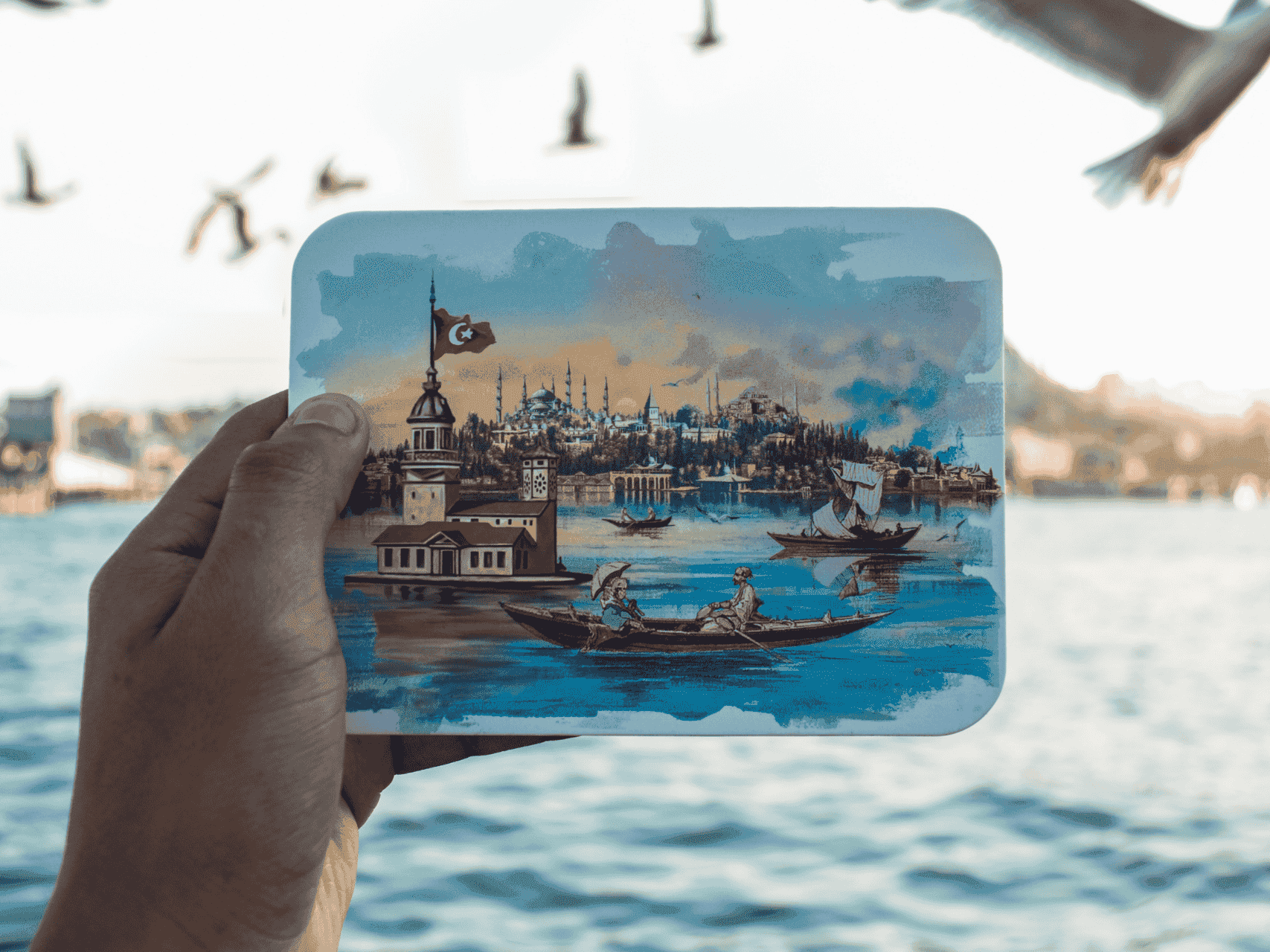 Mano sosteniendo una postal artística de Estambul con la Torre de la Doncella, Santa Sofía con gaviotas volando en el fondo.