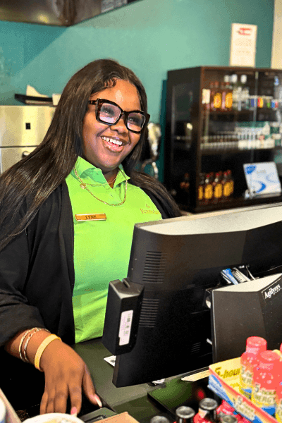 A young woman in glasses and a bright green shirt stands behind a register and smiles, an example of the exceptional customer service you'll find at Rosen Inn.