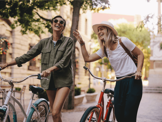 Two smiling women in casual attire with bicycles on a sunny street promoting Stay Longer, Save More offer.
