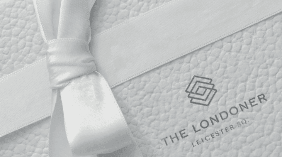 Close-up of a white gift box with a white ribbon, showcasing the logo of The Londoner Leicester Square