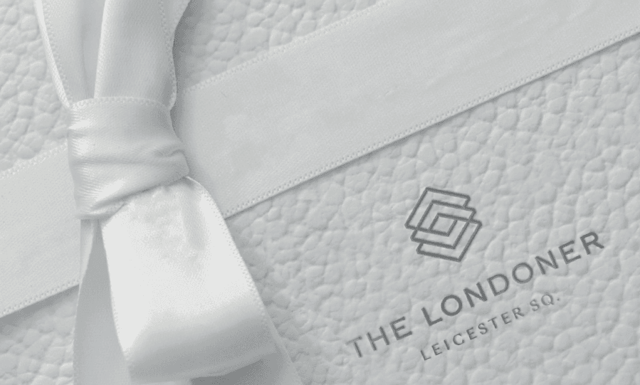 Close-up of a white gift box with a white ribbon, showcasing the logo of The Londoner Leicester Square