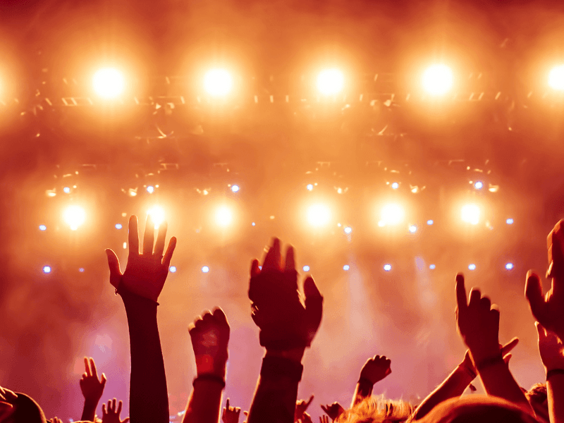 Concert crowd with raised hands silhouetted against bright orange stage lights and atmospheric smoke.