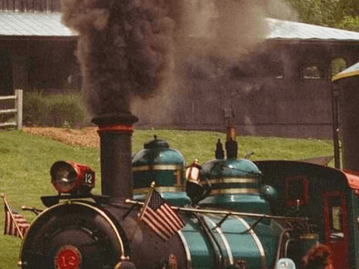 Steam locomotive number 12 emitting smoke with people nearby and a building in background.