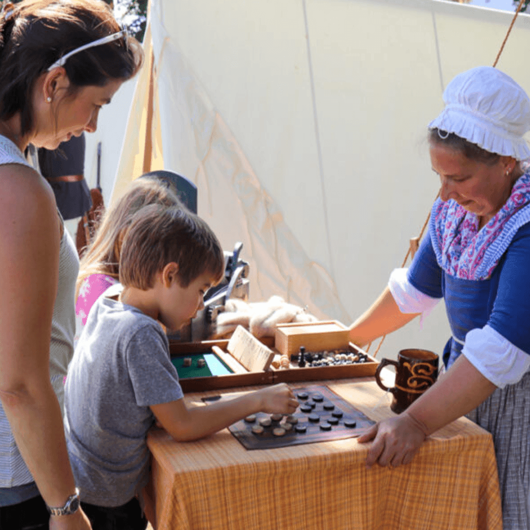 Woman in colonial attire guides children playing a board game on a table outdoors.