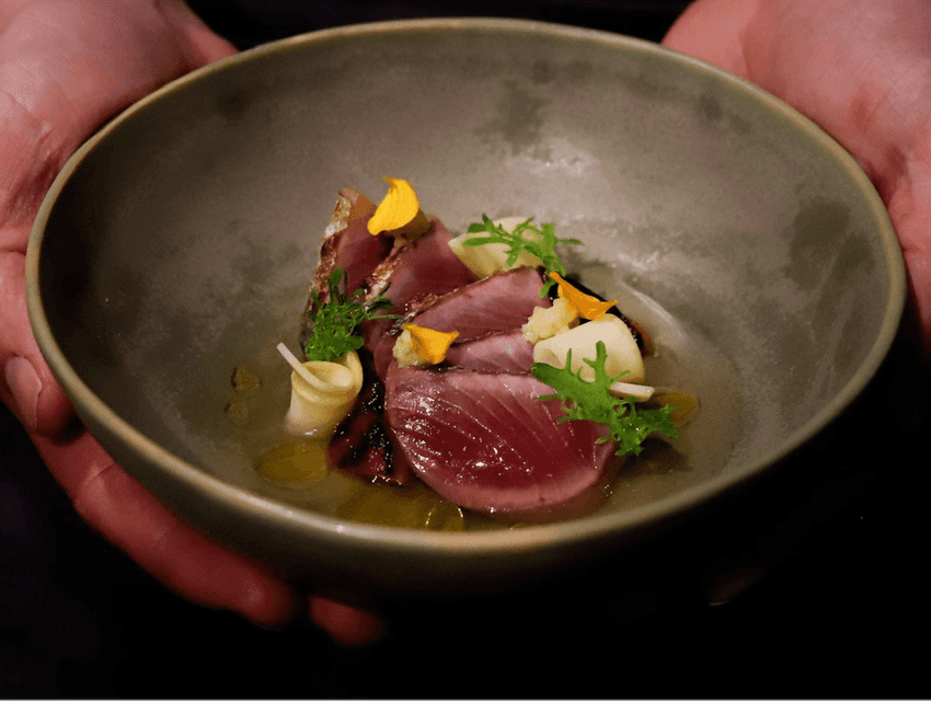 Hands holding a round bowl with a dish of sliced meat, herbs, and garnishes.