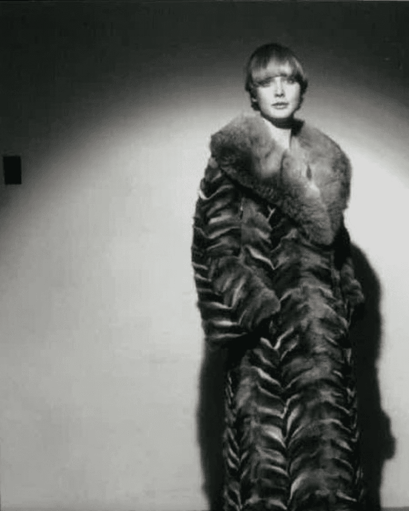 Black and white portrait of a woman in a long fur coat standing against a plain wall.