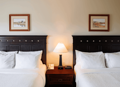 Two comfortable beds with white linens, dark wooden headboards & lamp in Deluxe Standard at Rodd Crowbush Golf & Beach Resort