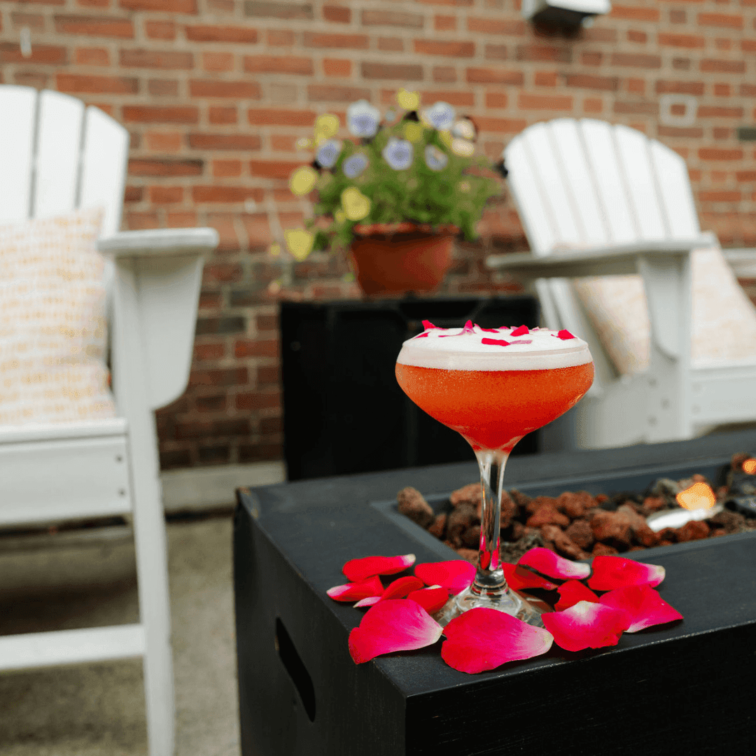 Cocktail with white and red petals on a black table by two white chairs and a brick wall.