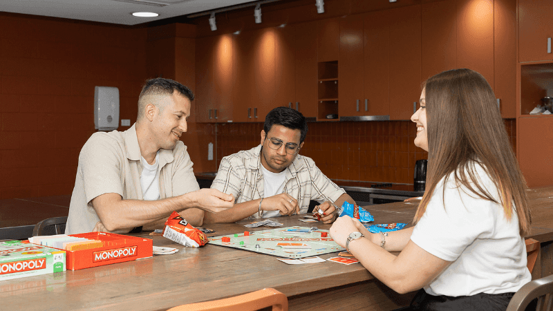 Three people playing Monopoly at a wooden table with snacks in a lounge.