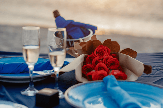 Two plates with blue napkins, two wine glasses, and a bouquet of red roses on a blue tablecloth.