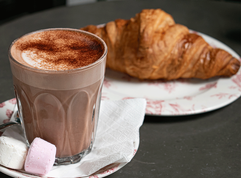 A hot chocolate with marshmallows with a croissant in the foreground
